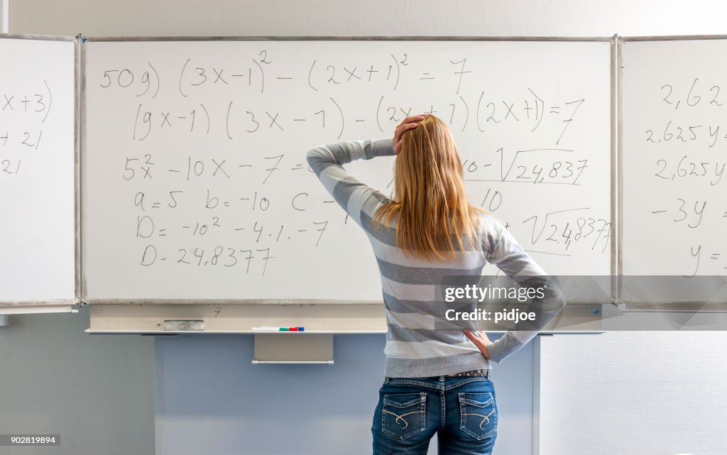 Drei Viertel Länge Schuss eines verwirrten und nachdenklich blonden High-School-Mädchen mit der Hand auf dem Kinn vor Whiteboard steht mathematische Formel Blick auf Brett