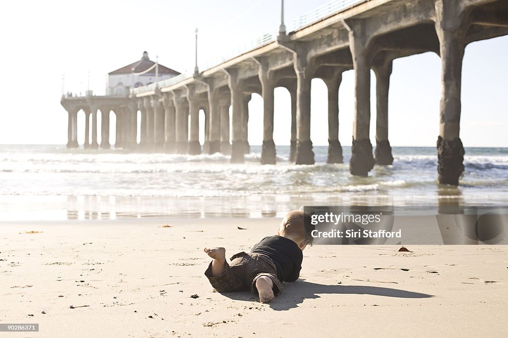 Young boy playing in sand at ocean, pier behind