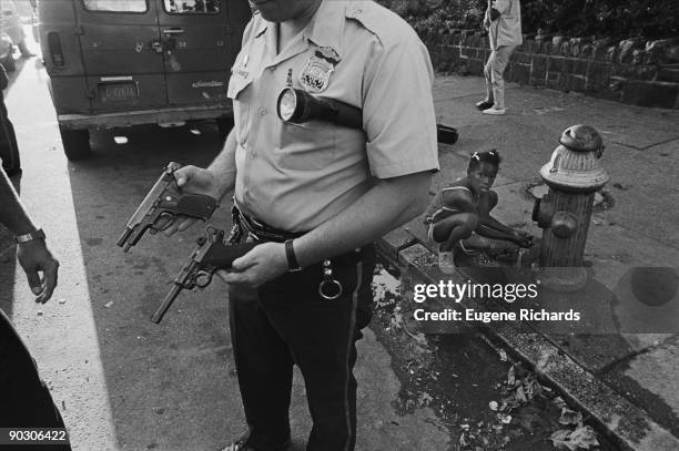 Police officer, with a flashlight under his arm, as he holds a pair of handguns that have been confiscated from a nearby house, North Philadelphia,...