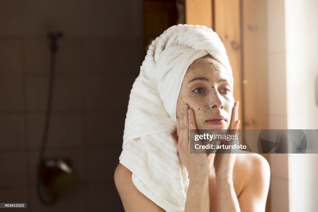Happy young woman applying face mask in bathroom. Beautiful female in front of mirror doing beauty treatment.