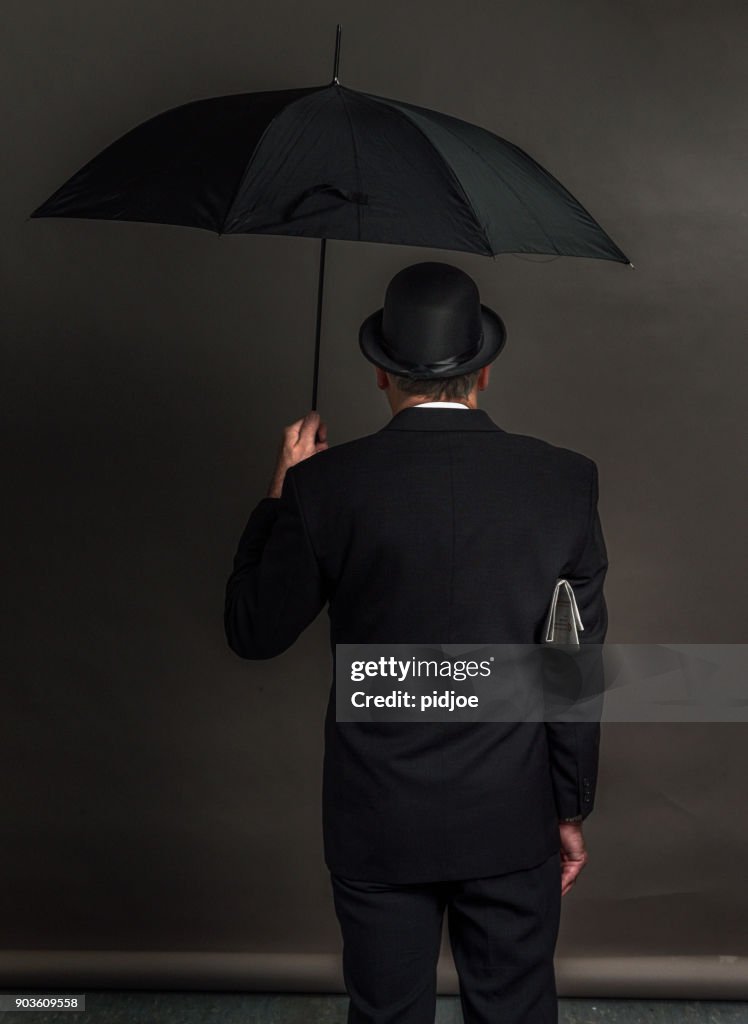 Rear view of an Englishman with bowler hat, umbrella and newspaper under the arm, grey background. Brexit concept: representing the choice in the British referendum on EU membership.