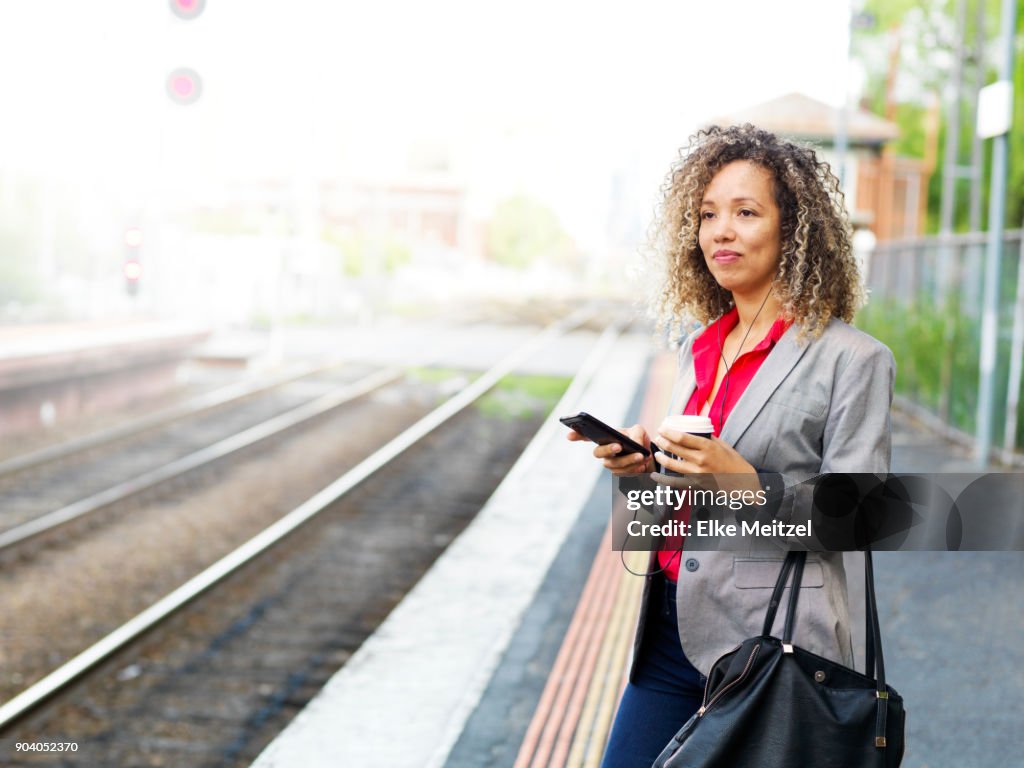 Woman with headphones, coffee cup and phone standing on train platform