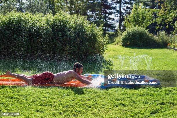 man playing on a slip and slide in the garden - backyard water slide stock pictures, royalty-free photos & images