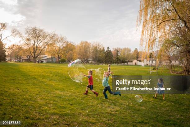four children chasing giant soap bubbles in a public park - public park kids stock pictures, royalty-free photos & images