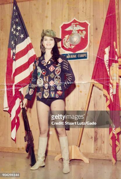Young, female American USO performer wearing suggestive clothing and a cowboy hat and boots, with a jacket which has many colorful patches, smiles...