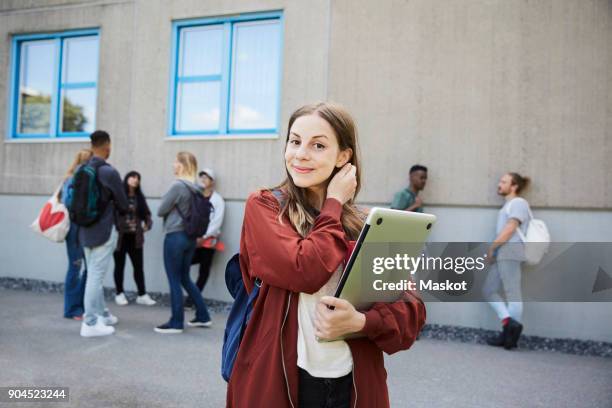 portrait of smiling female student carrying laptop while standing at university campus with friends in background - student laptop stock-fotos und bilder