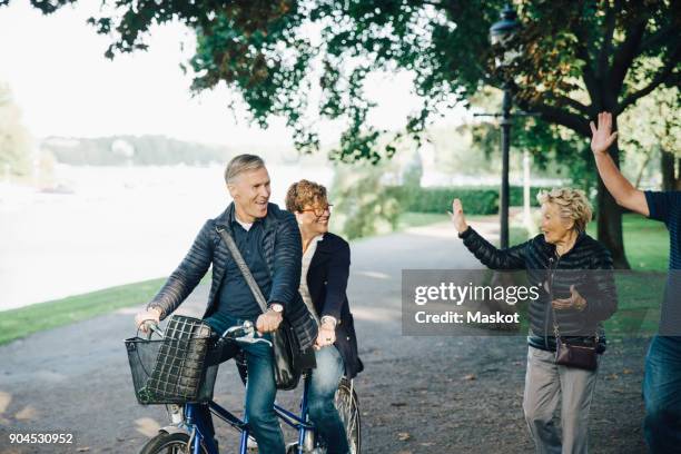 senior couple waving friends riding tandem bike in park - bicycle tandem stock-fotos und bilder