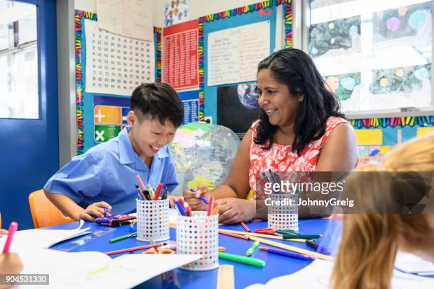 aboriginal school teacher smiling towards young asian boy working in classroom - native korean stock pictures, royalty-free photos & images