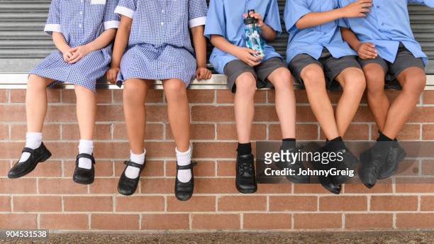 vista a bassa sezione di cinque bambini delle scuole seduti sul muro di mattoni indossando l'uniforme scolastica - calzature foto e immagini stock