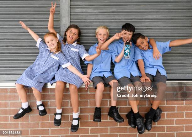 five school friends sitting on brick wall pulling faces and smiling towards camera - primary age child stock pictures, royalty-free photos & images