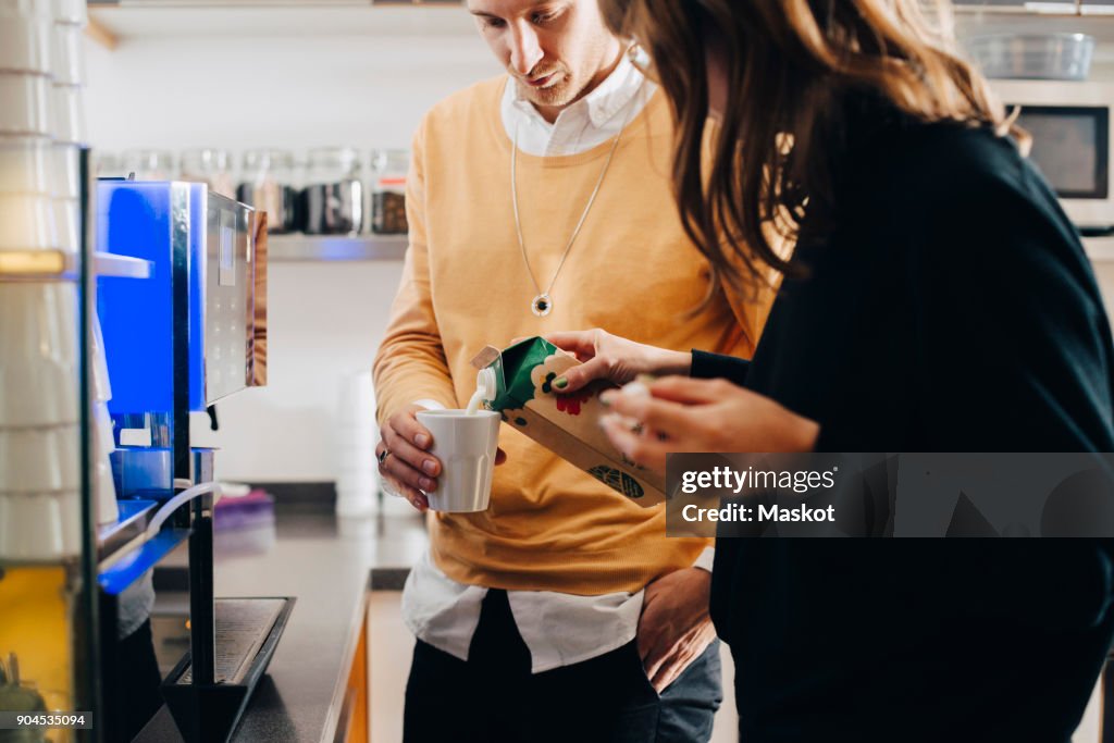 Midsection of businesswoman pouring milk in glass from carton for businessman at office cafeteria