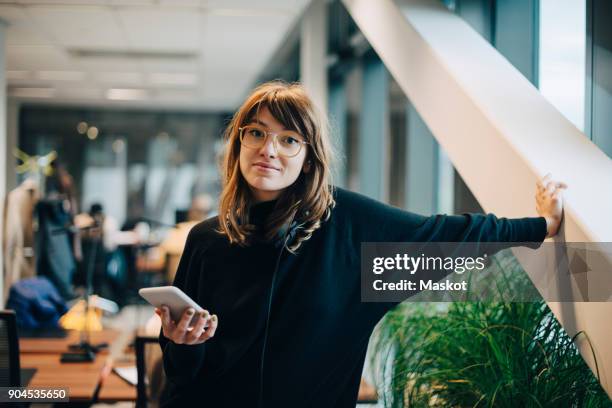 portrait of confident businesswoman standing with smart phone in office - business smartphone stockfoto's en -beelden