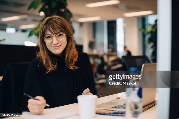 portrait of confident young businesswoman holding pen while sitting at desk in office - woman business desk front laptop office fotografías e imágenes de stock