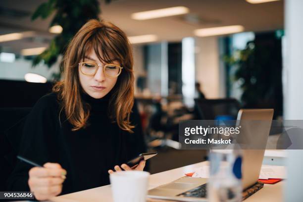 businesswoman writing while holding mobile phone at desk in office - mobile worker stockfoto's en -beelden