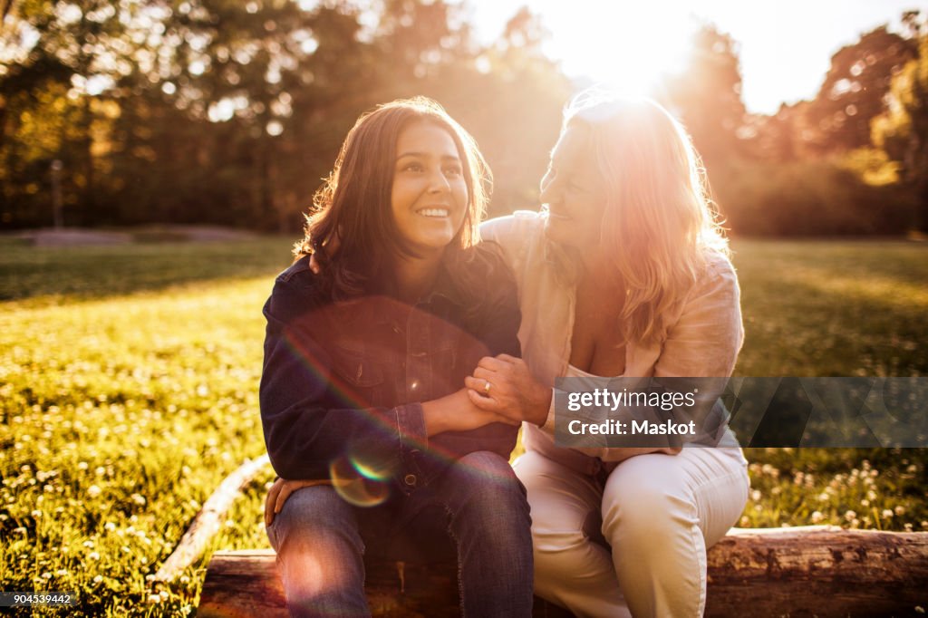 Mother looking at smiling daughter while sitting on log at park during sunny day