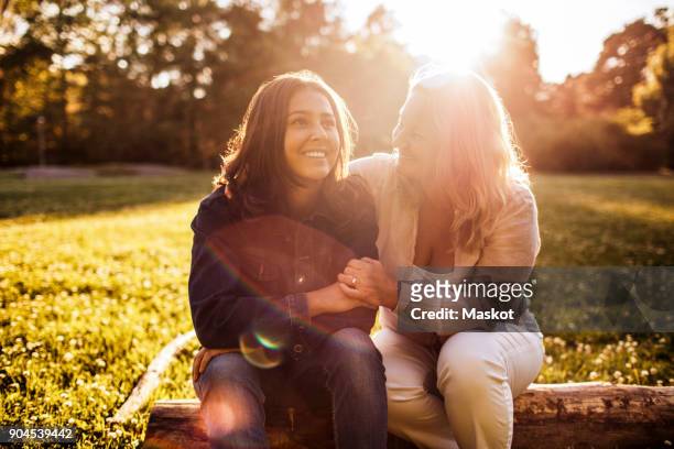 mother looking at smiling daughter while sitting on log at park during sunny day - family sadness stock pictures, royalty-free photos & images