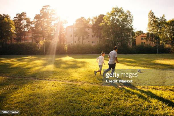 full length of father and son playing soccer on grassy field during sunny day - two guys playing soccer stockfoto's en -beelden