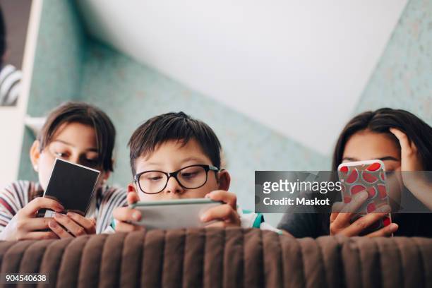 Low angle view of boy looking at digital tablet while lying with sisters using mobile phone on bed at home