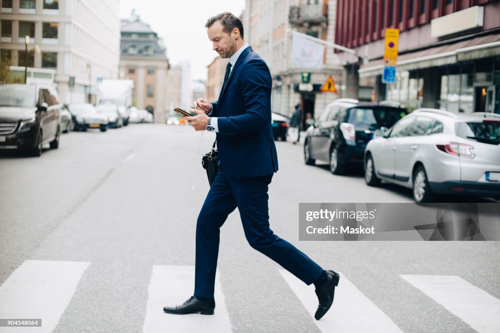 Full length side view of mature businessman crossing street while using smart phone in city