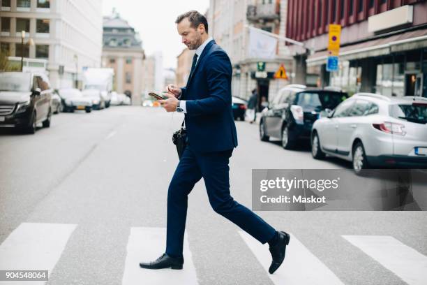 full length side view of mature businessman crossing street while using smart phone in city - business man walk stockfoto's en -beelden