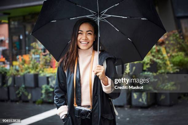 smiling woman with camera carrying umbrella while standing in city - female fashion with umbrella stock pictures, royalty-free photos & images