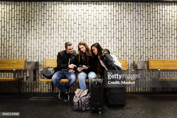full length of friends looking at smart phone while sitting against wall on subway station platform - metro platform stockfoto's en -beelden