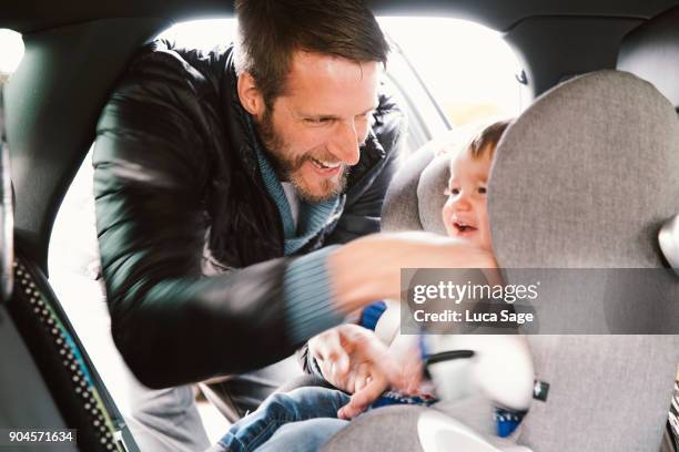 father laughs as he straps in his young toddler boy into his car seat - guy in car seat stockfoto's en -beelden