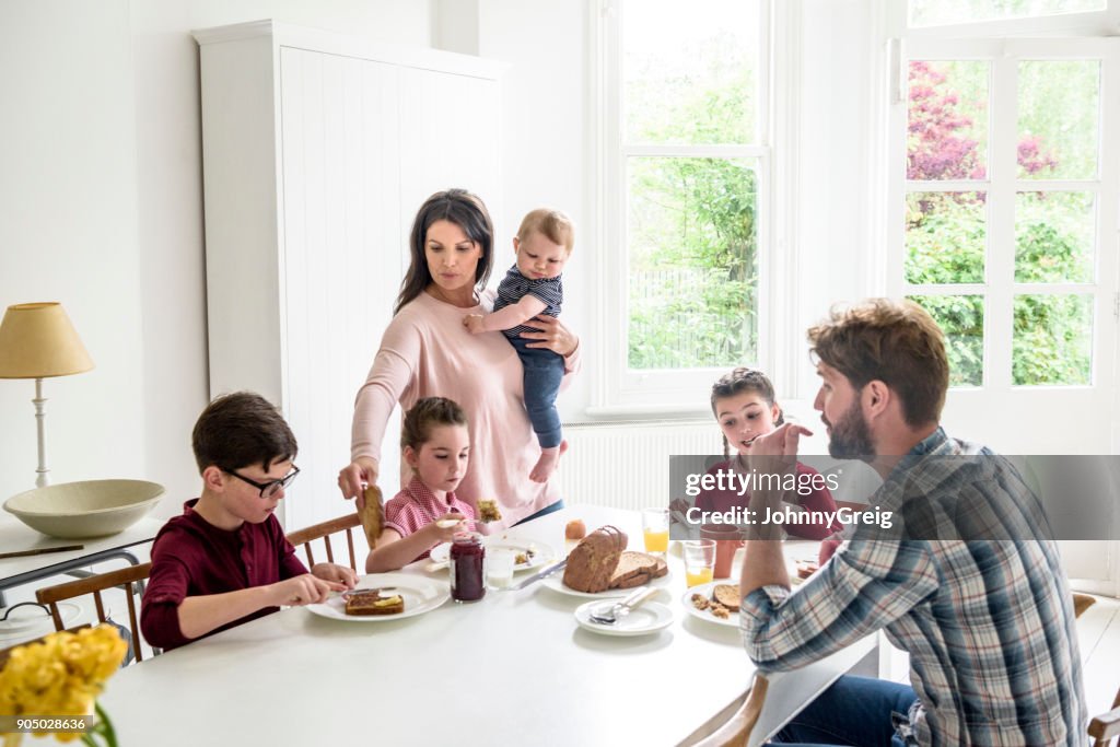 Família ocupada tomando café da manhã, a mãe serve torradas filho enquanto carregando menino