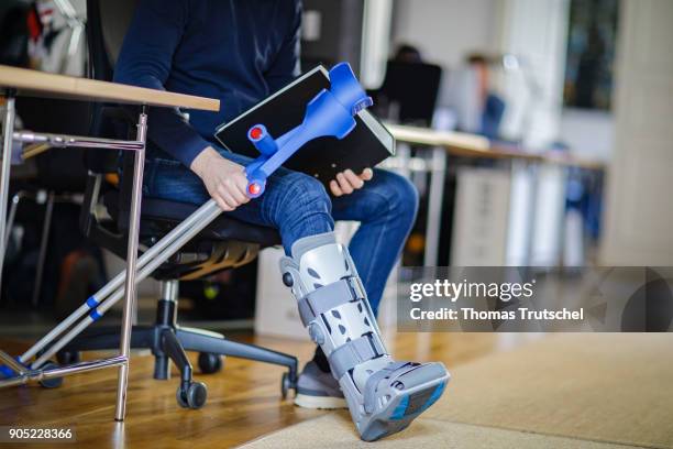 Berlin, Germany Symbolic photo on the topic: Sick at work. A man with crutches and an orthosis on his leg sitting at a desk in the office on January...