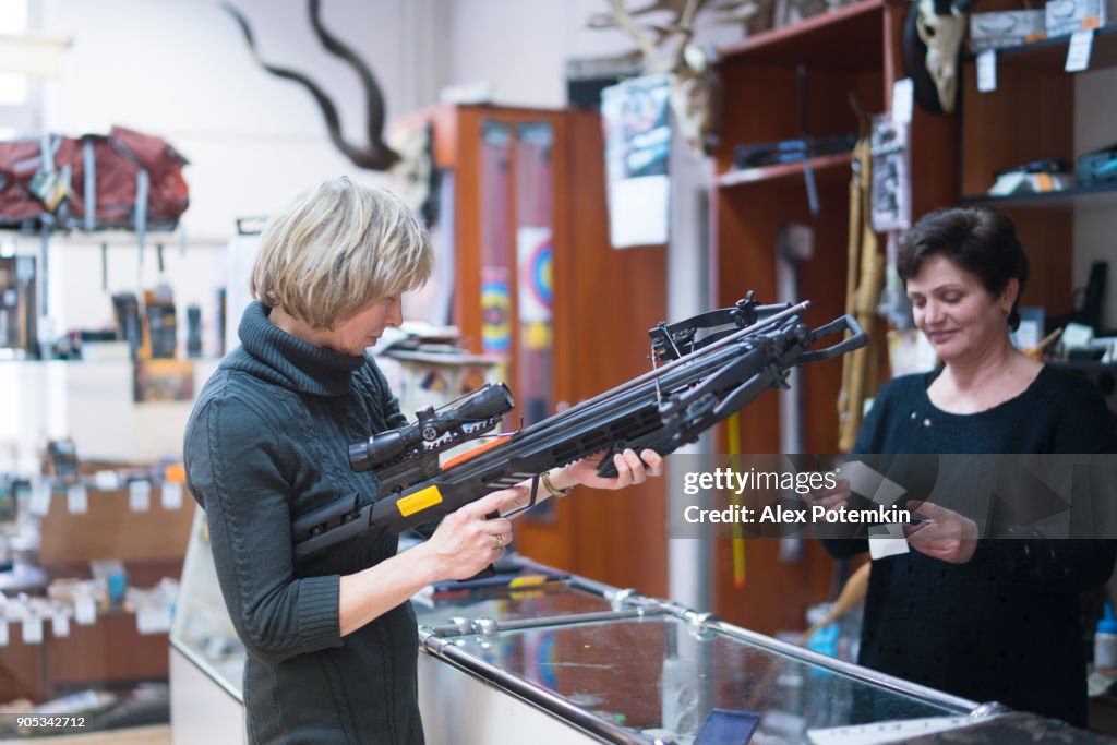 A loira madura, 50 anos de idade mulher atraente examinando o crossbow na loja de caça pequena, com a ajuda da mulher - vendas persone.