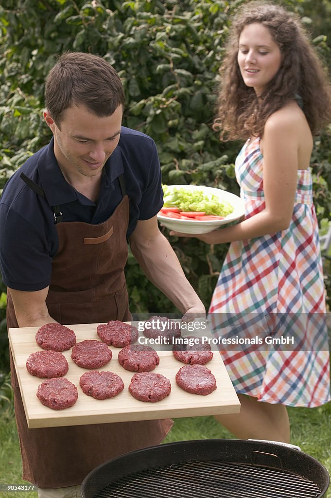 Man holding raw burgers over barbecue, woman holding salad