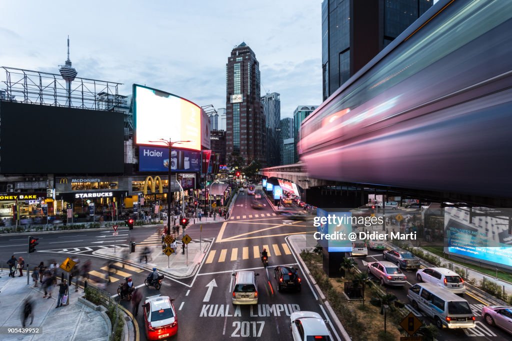 Traffic and a monorail car rush through the Bukit Bintang intersection at night in Kuala Lumpur