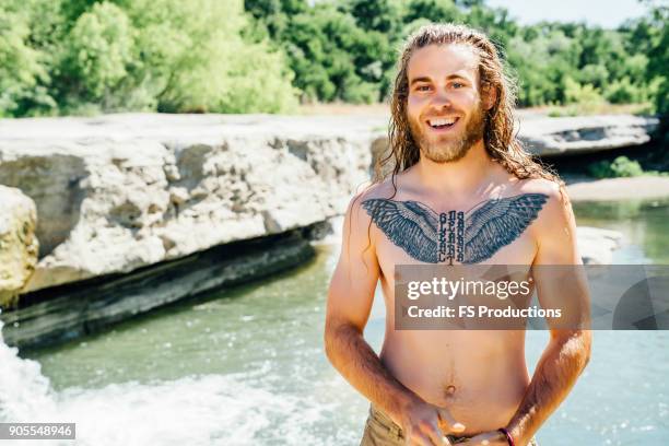 smiling caucasian man with chest tattoo standing near river - langer bart stock-fotos und bilder