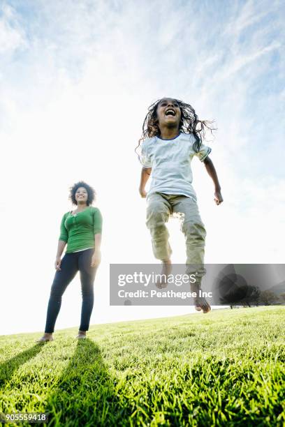 mixed race mother watching daughter jumping in the grass - afrikanisches kind beobachtet natur stock-fotos und bilder