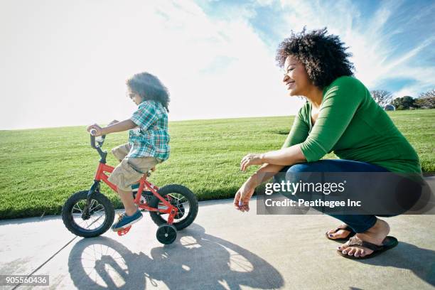 mixed race mother watching son ride bicycle - black woman riding bike foto e immagini stock