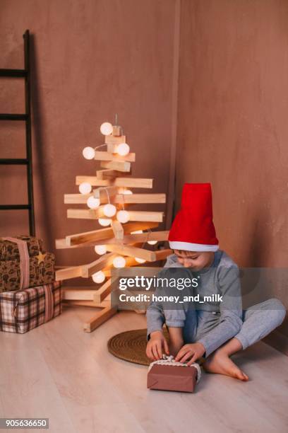 boy sitting on floor next to a modern christmas tree installation unwrapping a gift - honey boy fotografías e imágenes de stock