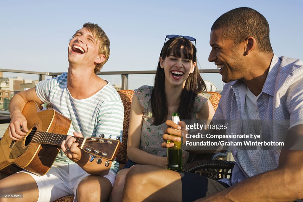 Three young adults laughing, one holding guitar