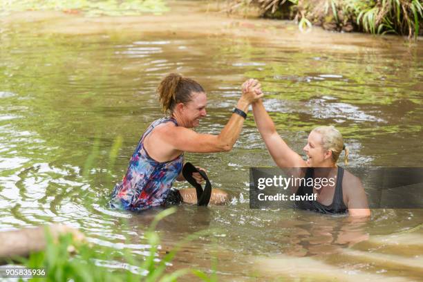 athletic mature women helping each other in a river during an obstacle course mud run - encouragement stock pictures, royalty-free photos & images