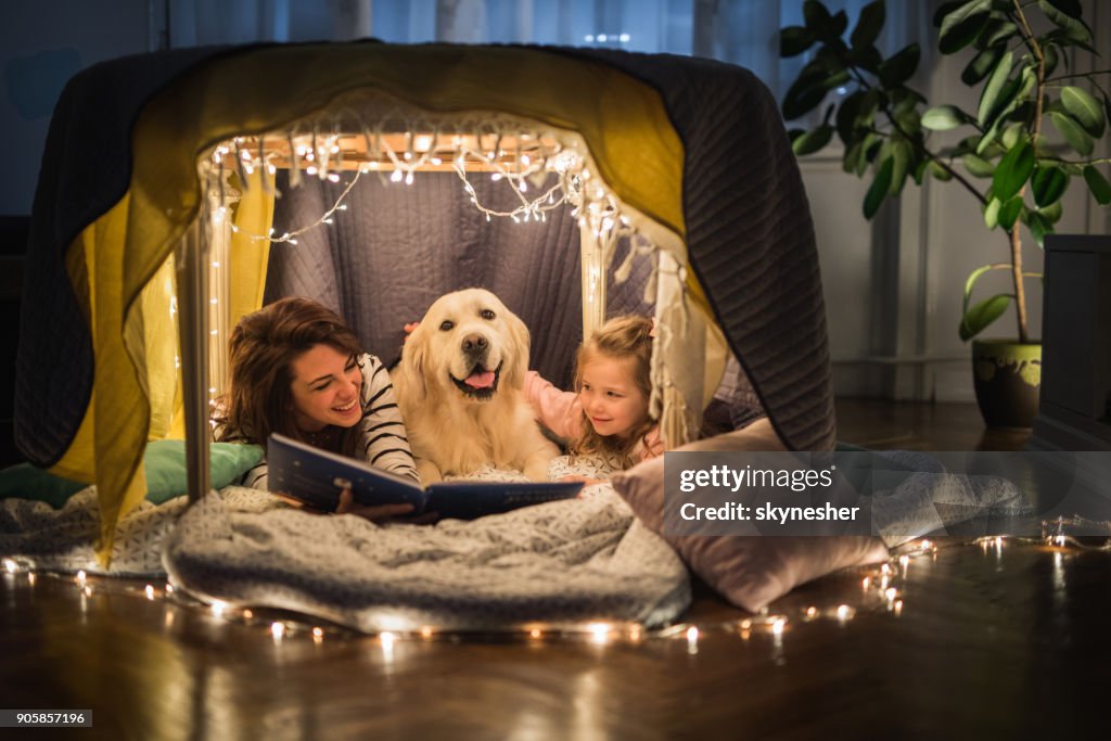 Gelukkig moeder en dochter ontspannen met een hond in een tent en lezen van een boek.