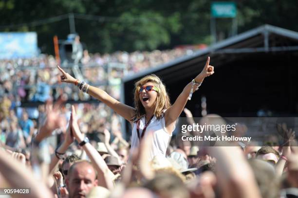 Young woman with her arms raised, smiling and cheering, sits on shoulders above the crowd on the second day of V Festival at Hylands Park on August...