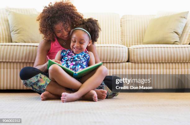 mixed race mother and daughter reading book on floor near sofa - espacio pequeño fotografías e imágenes de stock