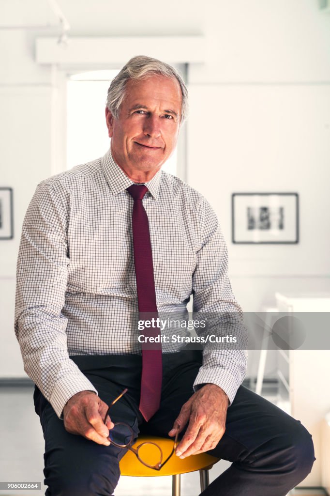 Smiling Caucasian businessman sitting on stool holding eyeglasses