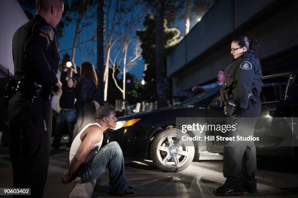 police standing around handcuffed man sitting on curb - police arrest stock-fotos und bilder