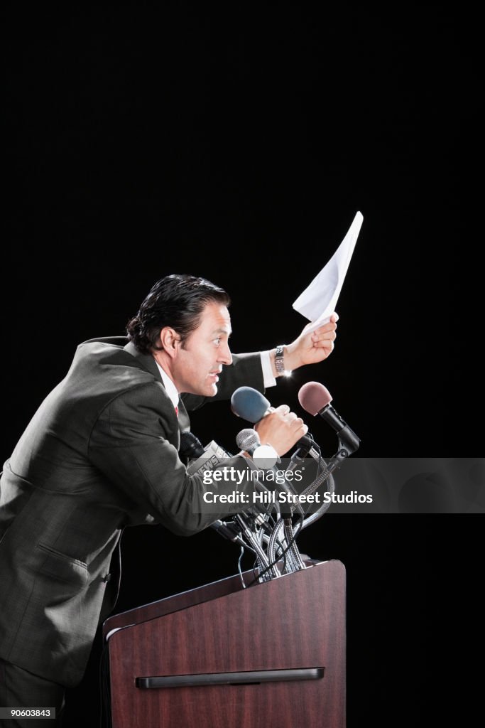 Hispanic man holding document at podium with microphones