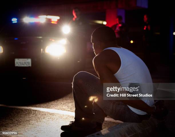 police shining lights on handcuffed african man sitting on curb - arresto foto e immagini stock