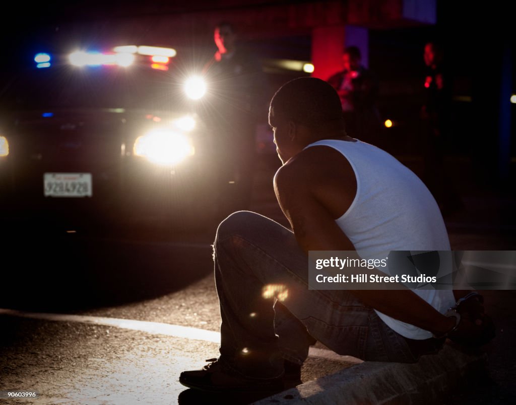 Police shining lights on handcuffed African man sitting on curb