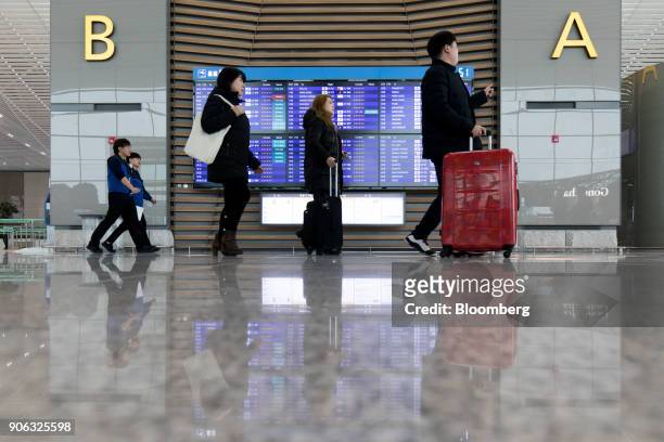 Travelers with luggage walk past screens showing flight departure information inside the terminal 2 building at Incheon International Airport in...