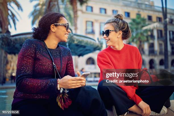 best girlfriends are texting and make fun sitting on the fountain - modern town square stock pictures, royalty-free photos & images