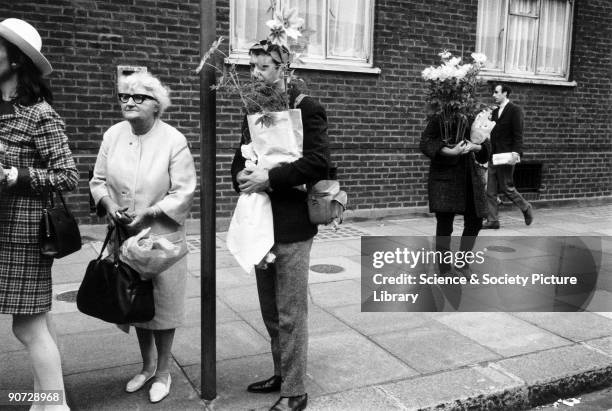 Young man and woman each holding a bouquet of flowers from Chelsea Flower Show, standing in a bus stop queue, 1968. Taken at Chelsea, London....