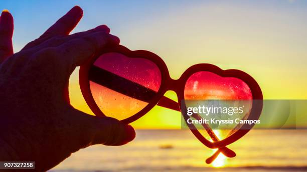 beautiful sunset through heart-shaped sunglasses on beach - hand gag stock-fotos und bilder